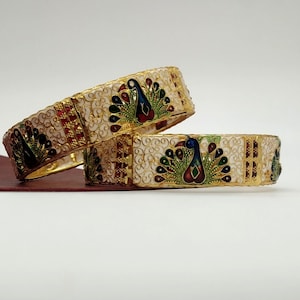 May include: Two gold bangles with white enamel and peacock designs. The peacocks are decorated with blue, green, red, and gold enamel.