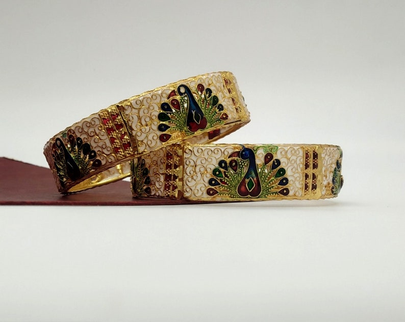 May include: Two gold bangles with white enamel and peacock designs. The peacocks are decorated with blue, green, red, and gold enamel.