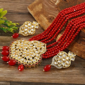 Puede incluir: Un collar de cuentas rojas con un colgante dorado y pendientes a juego. El colgante y los pendientes presentan un diseño floral con piedras blancas.