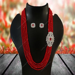 Puede incluir: Un collar de cuentas rojas y un juego de pendientes a juego exhibidos en un maniquí negro. El collar presenta múltiples hilos de cuentas rojas, una pieza central decorativa de color plateado con una piedra roja y pendientes cuadrados a juego.