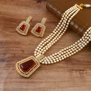 Op de afbeelding: Een gouden ketting met een rode edelsteen hanger en bijpassende oorbellen. De ketting is gemaakt van vijf strengen witte parels.