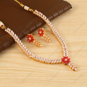 May include: A gold-toned necklace and earring set featuring white pearl-like beads and red flower accents. The necklace has a floral pendant and the earrings have a matching floral design. The jewelry set is displayed on a wooden surface.