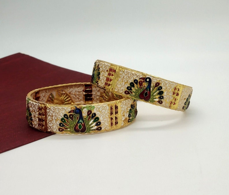 May include: Two white bangle bracelets with gold accents and peacock designs. The peacocks are decorated with blue, green, red, and gold enamel.