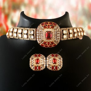 May include: A gold-toned jewelry set featuring a choker necklace and matching earrings. The necklace and earrings have a square design with a large red center stone, surrounded by smaller red and clear stones. The set is displayed on a black mannequin head.