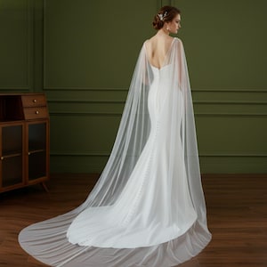 May include: A white wedding dress with a long train and a sheer cape. The dress has a fitted bodice and a row of buttons down the back. The cape drapes over the shoulders and extends to the floor.