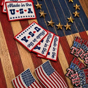 May include: An assortment of patriotic items, including patches with "Made in the USA" text, American flag patches, and gold star-shaped embellishments. The items are arranged on a wooden surface with red, white, and blue stripes.
