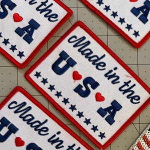 May include: White rectangular patches with a red border, embroidered with "Made in the USA" in navy blue. The letters "U", "S", and "A" are prominent, with red heart accents and star embellishments.