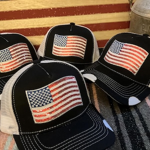 May include: Four black trucker hats with white mesh backs. Each hat features a sequined American flag design in red, white, and blue. The hats are displayed against a backdrop of a wooden American flag and a metal container.