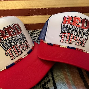 May include: Two red, white, and blue trucker hats with the text "RED WHITE TIPSY" in a patriotic design. The hats have a white front panel, a red brim, and a blue mesh back. The brim is decorated with rhinestones.