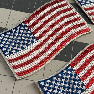 May include: Patches in the shape of the American flag. The patches feature a blue field with white stars and red and white stripes. The edges are trimmed with silver glitter. The patches are likely for clothing or accessories.