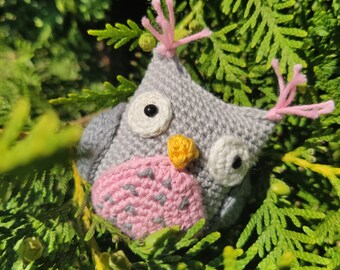 May include: A small, gray crocheted owl toy featuring a pink belly with gray stitched accents, white circular eyes with black pupils, a yellow beak, and pink yarn tufts on top of its head. The owl is crafted with a dense, textured yarn stitch.