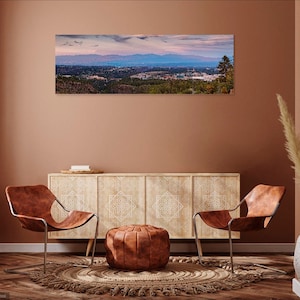May include: A panoramic photograph of a city skyline with mountains in the background. The photo is displayed on a wall in a living room setting with two chairs, a coffee table, and a vase with dried flowers.