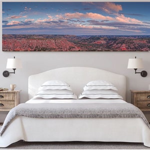 May include: A panoramic landscape photograph of a canyon with a cloudy sky. The photograph is hanging above a white bed with a gray blanket and two white pillows with black stripes. There are two wooden nightstands with lamps on either side of the bed.
