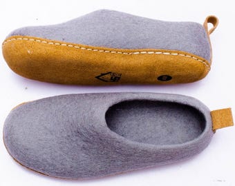 Puede incluir: Dos zapatillas de fieltro gris con suelas de cuero marrón y una pequeña presilla de cuero en el talón. Las suelas están cosidas a las partes superiores de fieltro gris. Las zapatillas se muestran desde dos ángulos diferentes.