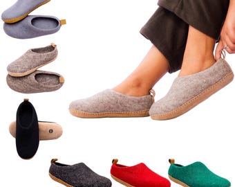 May include: A collection of felted wool slippers in various colors including light gray, brown, black, red, and green. Each slipper features a rounded toe, a tan leather-colored sole, and a small tan pull tab at the heel. The slippers have a simple, slip-on design with a soft, textured wool exterior.