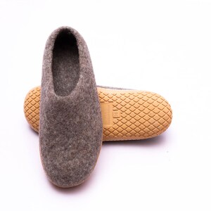 May include: A pair of gray felt slippers with a light brown rubber sole. One slipper is upright, showcasing the open top, while the other lies on its side, revealing the textured sole. The slippers are designed for indoor use.
