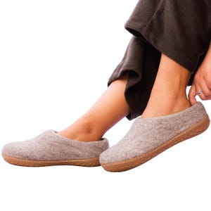 May include: A pair of light gray felt slippers with brown soles. The slippers are being worn by a person wearing dark brown pants. The slippers appear to be comfortable and suitable for indoor wear.