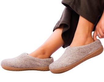 May include: A pair of light gray felt slippers with brown soles. The slippers are being worn by a person wearing dark brown pants. The slippers appear to be comfortable and suitable for indoor wear.