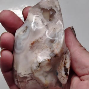May include: A polished flame-shaped stone carving featuring translucent white and pale gray agate with intricate banding, internal crystalline vugs, and small patches of earthy brown inclusions throughout the smooth, glossy surface.