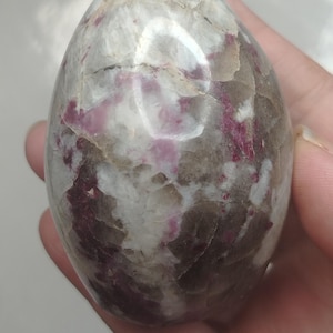 May include: A polished, egg-shaped stone featuring a marbled pattern of deep magenta, translucent grey, and opaque white mineral inclusions. The surface is smooth and reflective, showcasing natural crystalline fractures and varied colour veins.