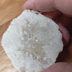 May include: A white crystal cluster with a rough, irregular shape, featuring clear quartz crystals at its core. The natural stone is held in a hand, highlighting its texture and colour variations.