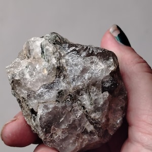 May include: A raw, unpolished mineral specimen featuring a cluster of translucent, clear, and smoky quartz crystals. The stone displays an irregular, jagged surface with visible internal fractures, inclusions, and dark mineral deposits embedded within the crystalline structure.