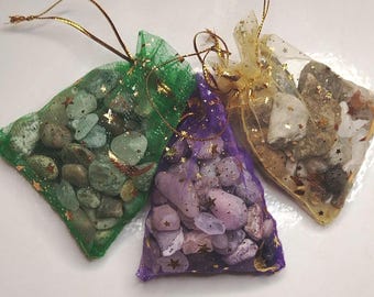 Puede incluir: Tres bolsas de organza transparente con cordón rellenas de piedras pulidas. Una bolsa verde contiene piedras verdes y grises, una bolsa morada contiene piedras de color lila y una bolsa dorada contiene piedras beige, grises y blancas.