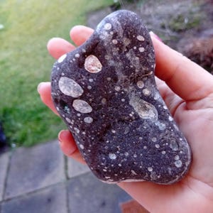 Könnte beinhalten: Ein dunkelgrauer Stein mit weißen und beigen Flecken. Der Stein wird in einer Hand gehalten und hat eine glatte Oberfläche. Der Stein hat eine einzigartige Form und ist wahrscheinlich ein Naturstein.