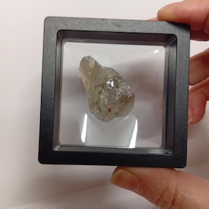 Può includere: Un cristallo grezzo, di colore chiaro, è esposto all'interno di una teca quadrata nera. Il cristallo ha una forma naturale e irregolare e appare traslucido. La teca è tenuta da una mano su sfondo bianco.