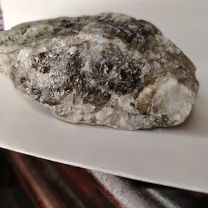 May include: A raw, unpolished mineral specimen featuring a mix of translucent smoky gray and opaque white crystalline structures. The rock has an irregular, jagged surface with visible facets and varying textures throughout its entire form.