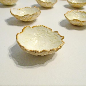 Könnte beinhalten: Eine Sammlung kleiner, dekorativer Schalen mit einer einzigartigen, organischen Form. Die Schalen haben einen cremeweißen Innenraum und einen strukturierten, goldfarbenen Rand. Sie sind auf einer weißen Oberfläche angeordnet.