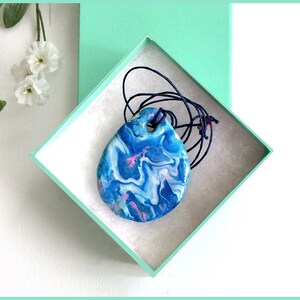 May include: A teardrop-shaped pendant with a blue, white, and pink marbled design, strung on a dark blue cord. The pendant is displayed inside a light green box with a white interior, set against a white background.