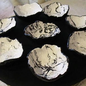 Può includere: Un set di otto ciotole in ceramica fatte a mano. Le ciotole sono bianche con marmorizzazione nera e bordi neri, disposte su una base circolare nera. Ideali per servire snack o piccoli piatti.