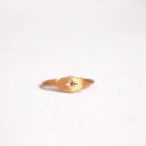 May include: A gold ring featuring a small pink gemstone set in a star-shaped design. The ring is oval-shaped and has a textured surface.