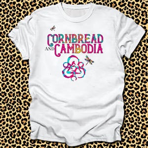 Può includere: T-shirt bianca con la scritta "CORNBREAD AND CAMBODIA" in lettere colorate e stilizzate. Il design include illustrazioni di libellule e una grafica astratta a spirale nei toni del blu, rosa e giallo.