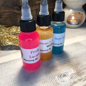 May include: Three small glass bottles with black caps and clear droppers filled with pink, yellow, and blue liquids. The bottles have labels with the text "Fruit Loops Warming Oil", "Pecan Puff Waffle Warming Oil", and "High Tide Warming Oil".