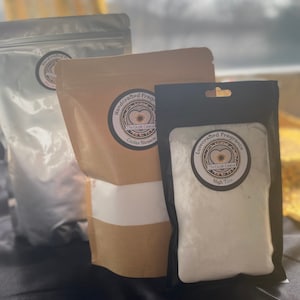 Könnte beinhalten: Drei weiße Beutel mit handgemachtem Duft, zwei in braunen Papiertüten mit schwarzen Etiketten und einer in einer schwarzen Tüte mit einem weißen Etikett. Die Etiketten haben ein Sonnenblumendesign und den Text "The Crack Caramel" und "Handgemachter Duft". Der erste Beutel sagt "Cactus Blossom" und der zweite "High Tide".