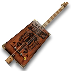 Puede incluir: Una guitarra de caja de puros de madera con un diseño de la Ruta 66 y el texto "American Road 1926 U.S.A." y "Free Soul".