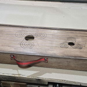 May include: A wooden musical instrument with a rectangular body and a handle. The instrument has strings and a decorative design on the top.