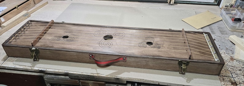May include: A wooden musical instrument with a rectangular body and a handle. The instrument has strings and a decorative design on the top.