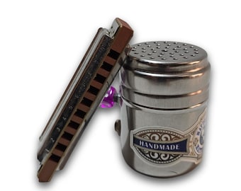 May include: A silver-colored harmonica leans against a matching metal shaker with a perforated top. The shaker has a label that reads "HANDMADE". A purple accent is visible.