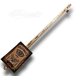 May include: A wooden cigar box guitar with a brown wooden body and a natural wood neck. The body is decorated with a label that reads "Mattacacci's" in white lettering.