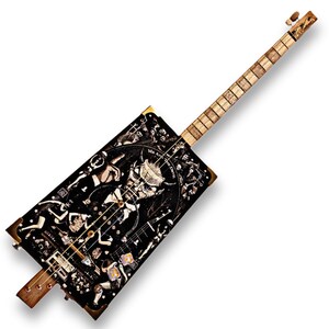 Puede incluir: Una guitarra de caja de puros negra y dorada con una ilustración detallada de una criatura con cuernos y el texto "Mr. A.C. The Devil". La guitarra tiene un mástil de madera y cuerdas.