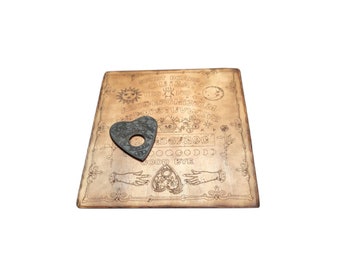 May include: A wooden Ouija board with a planchette in the shape of a heart. The board has a brown background with black lettering and symbols. The planchette is black with a silver trim.