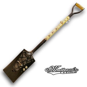 May include: A black shovel guitar with a white and black patterned design on the shovel head. The handle is wrapped in a cream-colored material with a black design. The guitar has a black pickguard with a white dice design. The text "Matteacci's Guitars" is visible in the lower right corner of the image.