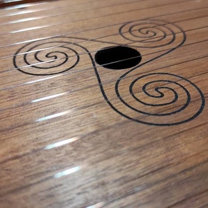 May include: A wooden musical instrument with a sound hole and a Celtic knot design. The instrument has a light brown finish and is made of wood.