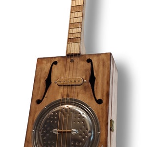 May include: A wooden cigar box guitar with a metal resonator, a pickup, and a wooden neck. The guitar has a brown stain and a distressed look.