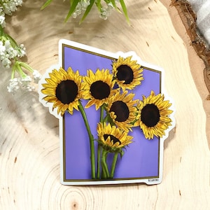 Può includere: Un adesivo con girasoli con petali gialli e centri marrone scuro, su uno sfondo viola chiaro. L'adesivo ha un bordo bianco e steli verdi. L'adesivo è su una superficie di legno.