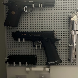 Può includere: Un assortimento di armi da fuoco, tra cui due pistole nere e un revolver argentato, montate su una pannello forato grigio. Le pistole hanno impugnature nere e dettagli argentati. Il revolver ha un'impugnatura nera e una canna lunga.