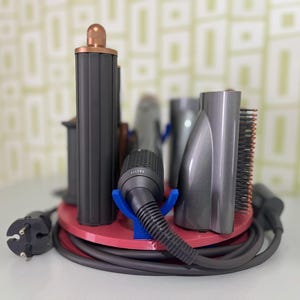 May include: A hair styling tool organizer with various attachments. The organizer is a red circular base with a black power cord. The attachments are gray and black, including a brush, nozzle, and a cylindrical piece with a copper-colored top.